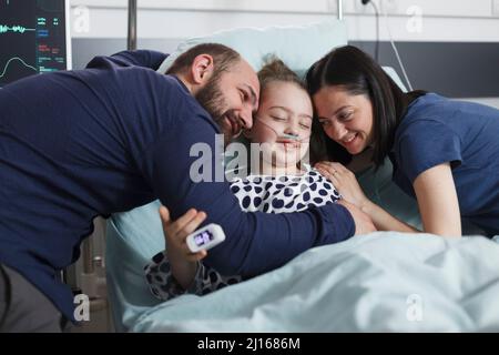 Fröhliche, glückliche Eltern sitzen zusammen mit einem lachenden kleinen Mädchen, während sie sich im Zimmer der Krankenhausinternationstation umarmen. Die kranke Tochter ist positiv und fröhlich, während Mutter und Vater sie trösten. Stockfoto