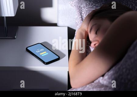 Der Wecker weckt müde schlafende Frau nachts oder morgens im Bett. Mobiltelefon auf dem Tisch mit Zeitschaltuhr und Schlummertaste. Überschlafende Person. Stockfoto