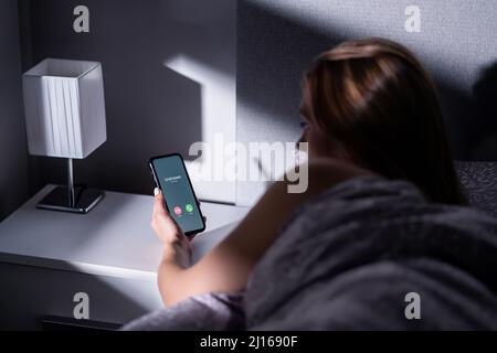 Telefonbetrug von unbekanntem Anrufer. Smartphone-Betrug durch Handynummer in der Nacht. Frau im Bett antwortet auf ankommende Schwindel, Streich oder Phishing-Verbrechen. Stockfoto