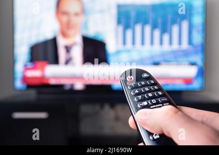 TV-Nachrichtenreporter auf Fernsehbildschirm und Fernbedienung. Mann, der Moderator, Anker oder Newsreader im Studio beobachtet. Newstcast-Kanal wird durchbrochen. Stockfoto