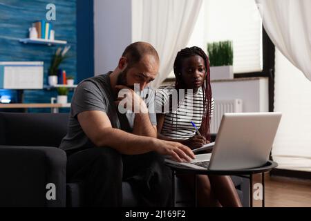 Besorgt multiethnischen Paar Blick auf Laptop-Bildschirm, während afroamerikanische Frau Notizen. Gestresste junge Erwachsene arbeiten zusammen, um falsche Bankrechnungen mithilfe von Online-Buchführung zu lösen Stockfoto
