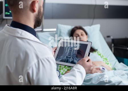 Medic hält ein digitales Tablet mit Hals-mrt und betrachtet den schlafenden Patienten im Krankenhausbett nach einer Rachenoperation. Frau, die nach einem chirurgischen Eingriff mit dem Bildschirm zur Messung der Vitalparameter verbunden ist. Stockfoto