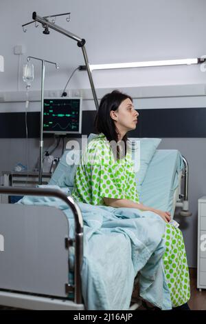 Gelangweilte Frau, die eine infusionsleitung erhalten hat und auf der Krankenstation auf dem Bett sitzt und in die Ferne blickt. Patient mit Krankheit fühlt sich depressiv und wartet auf den Arzt, um klinische Beratung zu tun. Stockfoto