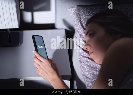 Schlaflosigkeit. Schlaflose Frau, die die Zeit nicht in der Nacht schlafen. Traurige müde Person mit Störung im Bett. Telefonuhr. Ein schlechter Traum. Stockfoto