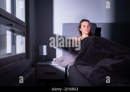Frau, die im Bett ein Handy benutzte, lächelte glücklich. Spät arbeiten oder SMS in der Nacht vor dem Schlafen schreiben. Smartphone-Chat oder SMS. Schlafzimmer zu Hause. Stockfoto