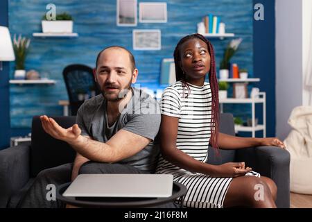 Multiethnisches Ehepaar, das sich auf dem Sofa im Wohnzimmer über finanzielle Instabilität streiten muss. Vereiterte Partner sitzen auf der Couch, während sie einen ernsten worying Konflikt haben Stockfoto