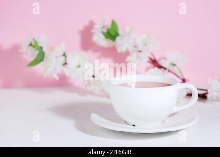 Weißer Becher mit rosa Tee, auf einer Untertasse, auf einem Betontisch, auf einem rosa Hintergrund mit Blumen. Harte Schatten. Die Getränke. Speicherplatz kopieren Stockfoto