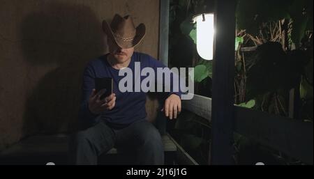 Cowboy-Mann kommuniziert online mit seinem Telefon, während er auf der Veranda sitzt. Nacht Stockfoto