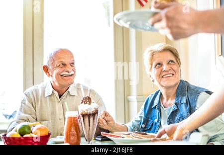 Der Kellner serviert ein Rentnerpaar, das im Fashion Bio Restaurant Kuchen isst - Pension und aktives Senioren-Konzept mit erwachsenen Menschen, die echten Spaß haben Stockfoto