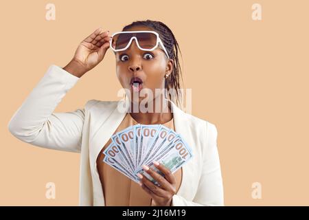 Junge Frau mit überraschendem Ausdruck, die mehrere hundert Dollar auf beigefarbenem Hintergrund hält. Stockfoto