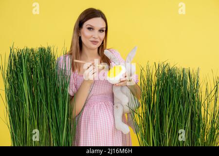 Junge süße Schwangerin trägt ein rosa, weißes Kleid mit beigefarbenem Spielzeug-Kaninchen und malt ein großes künstliches Ei. Stockfoto