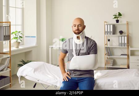 Trauriger Mann mit gebrochenem Arm in der Schlinge und einem Halskragen, der im Krankenhaus sitzt. Stockfoto