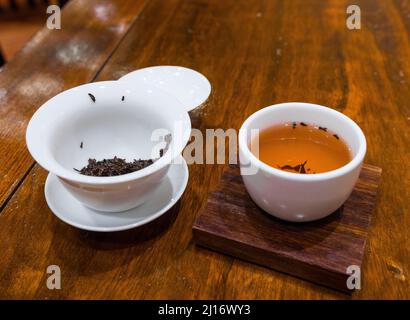 Gong Fu Cha brauen. Schönes weißes Porzellan (Gaibei/Gaiwan & Teetasse) auf einem Holztisch. Nasse rote Teeblätter. Perfekt für die Teeverkostung Stockfoto