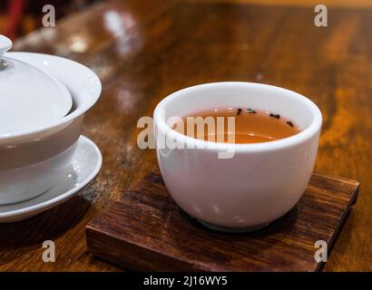 Gong Fu Cha brauen. Schönes weißes Porzellan (Gaibei/Gaiwan & Teetasse) auf einem Holztisch. Nasse rote Teeblätter. Perfekt für die Teeverkostung Stockfoto