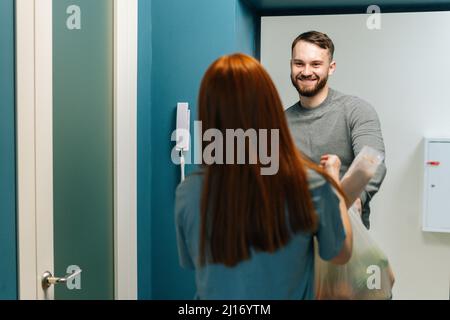 Lächelnder, bärtiger Kurier, der zu Hause am Eingang Lebensmittelbeutel an eine nicht erkennbare weibliche Hausfrau lieferte. Stockfoto