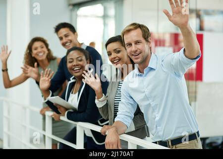 Hallo Erfolg. Porträt einer Gruppe lächelnder Mitarbeiter, die winken, während sie sich in einem Büro auf ein Geländer stützen. Stockfoto