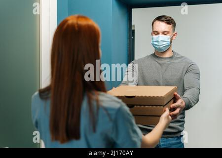 Kurier männlich trägt medizinische Gesichtsmaske liefert Boxen mit heißer Pizza zu unkenntlich weiblichen Kunden vor der Haustür zu Hause. Stockfoto