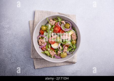 Draufsicht auf gesunden und leckeren Salat mit Thunfisch, frischer Rucola, Kirschtomaten, Wachtelei, Zwiebel und grünen Oliven auf grauem Betongrund Stockfoto