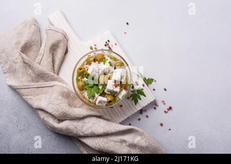 Draufsicht auf schmackhafte grüne Oliven mit Feta-Käse, Pfeffer und frischer Petersilie in einer Glasschüssel mit Öl auf grauem Betongrund Stockfoto