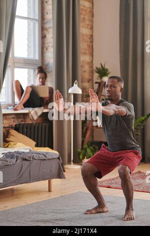 Vertikale Vollaufnahme des modernen jungen schwarzen Mannes, der eine stärkende Übung macht, während seine Freundin im Hintergrund im Internet surft Stockfoto