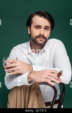 Mann mit Schnurrbart, der Zigarre und Glas Whiskey hält, während er sich auf einem dunkelgrünen Stuhl lehnt Stockfoto