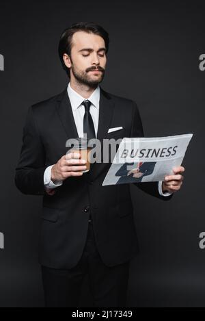Mann mit Schnurrbart, der einen Pappbecher hält und die Geschäftszeitung auf Dunkelgrau liest Stockfoto