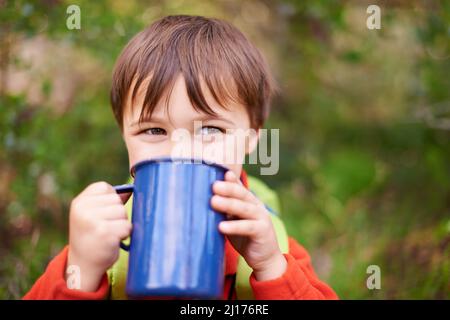 Vorbereitung auf einen energiegeladenen Campingtag. Aufnahme eines entzückenden kleinen Jungen, der während eines Campingausflugs aus einem Becher trinkt. Stockfoto