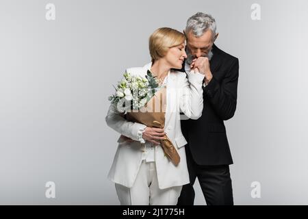 Reifer Bräutigam küsst Hand der Braut in Anzug hält Hochzeit Bouquet auf grau isoliert Stockfoto