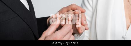 Beschnittene Ansicht des Bräutigams mit Ehering am Finger der Braut isoliert auf grauem Banner Stockfoto