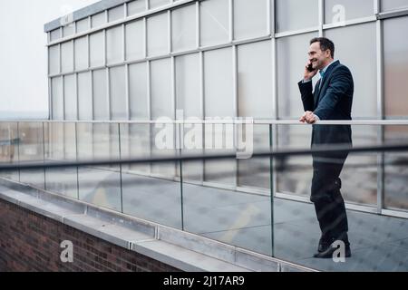 Lächelnder Geschäftsmann, der am Glasgeländer auf dem Balkon mit dem Smartphone spricht Stockfoto