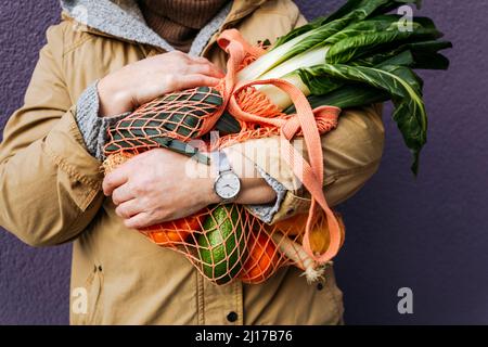 Frau, die einen Netzbeutel mit Gemüse vor der lila Wand hält Stockfoto