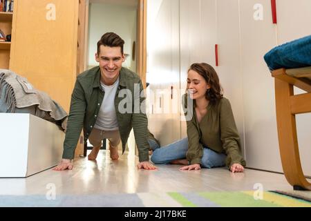 Glückliche junge Frau, die zu Hause einen Freund ansieht, der Liegestütze macht Stockfoto