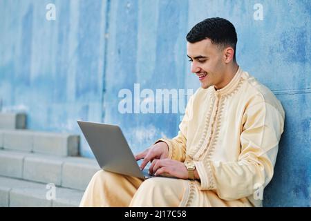Fröhlicher junger Mann mit Laptop, der auf einer Treppe an der blauen Wand sitzt Stockfoto