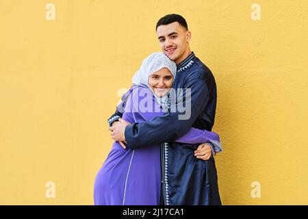 Lächelndes junges Paar, das sich umarmt und vor einer gelben Wand steht Stockfoto