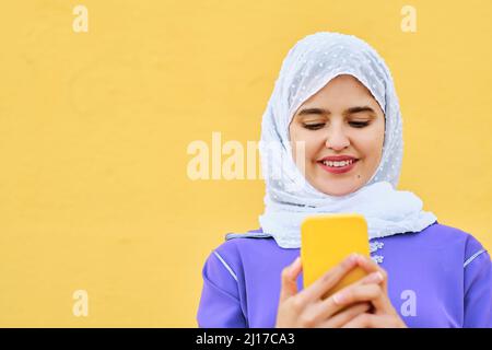 Lächelnde junge Frau, die vor der gelben Wand durch das Smartphone im Netz surft Stockfoto