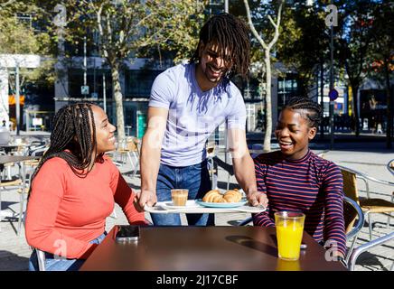 Glücklicher junger Mann, der Tablett für Freunde im Straßencafé auf dem Tisch trug Stockfoto