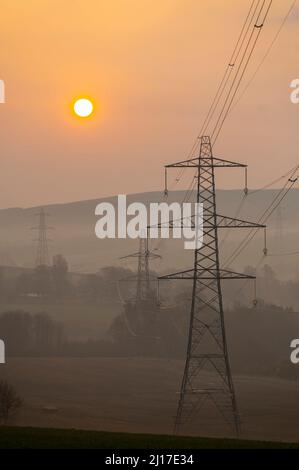 Ledgerwood in Scottish Borders, Großbritannien. 23.. März 2022. Energie, Strom, Strom. Die Sonne geht an einem anderen Tag mit hohen Energiepreisen auf, während die Lebenshaltungskosten weiter steigen. Der Übergang zu grüner Energie steht ganz oben auf der Tagesordnung, aber auch die Bilanz der Senkung der Energiekosten, die derzeit aufgrund der covid Pandemie und des aktuellen Kriegs in der Ukraine und der Sanktionen gegen Russland hoch ist. Der Blick ist auf Strommasten und eine ferne Windturbine, wenn die Sonne in der Nähe von Ledgerwood in den schottischen Grenzen aufgeht. Quelle: phil wilkinson/Alamy Live News Stockfoto