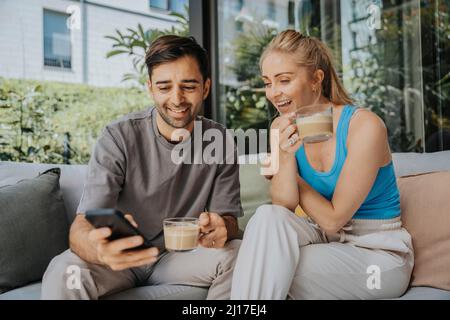 Mann, der in der Lounge ein Smartphone mit einer Frau teilt, die Kaffee auf dem Sofa getrunken hat Stockfoto