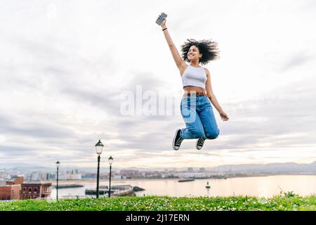 Fröhliche junge Frau mit Smartphone springen in der Natur Stockfoto