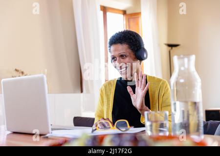 Lächelnde Geschäftsfrau mit Kopfhörern, die bei Videoanrufen über den Laptop zu Hause winken Stockfoto