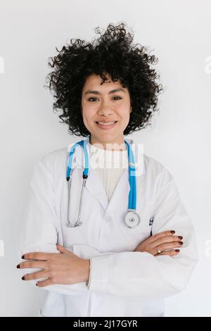 Glücklicher junger Arzt, der mit gekreuzten Armen vor einer weißen Wand steht Stockfoto
