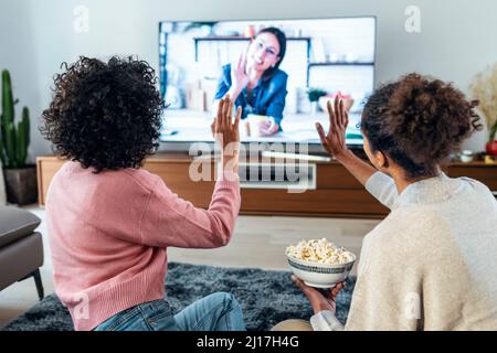 Frauen winken bei einem Videoanruf mit Freunden über das Fernsehgerät zu Hause mit den Händen Stockfoto