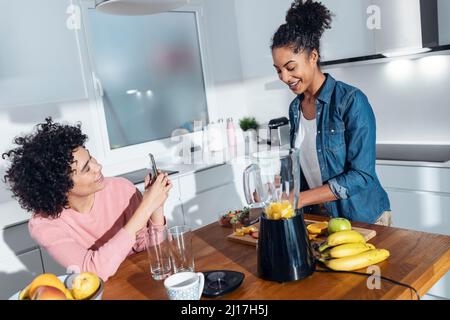 Lächelnde junge Frau fotografiert einen Freund, der in der Küche zu Hause Saft zubereitet Stockfoto