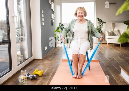 Lächelnde ältere Frau mit Widerstandsband sitzt auf einem Stuhl und arbeitet zu Hause Stockfoto