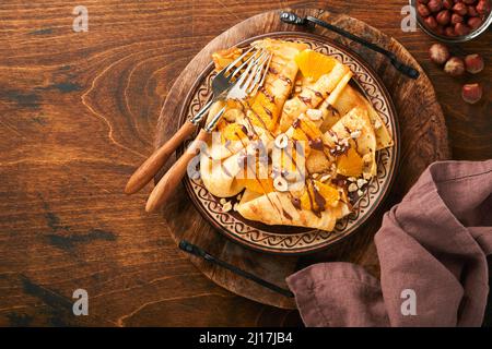 Crepes-Suzette mit Orangen und Schokolade. Dünne Crepes mit Schokoladenaufstrich, Haselnüssen und Orangenscheiben Obst auf dem Teller zum Frühstück auf altem dunklen Woo Stockfoto