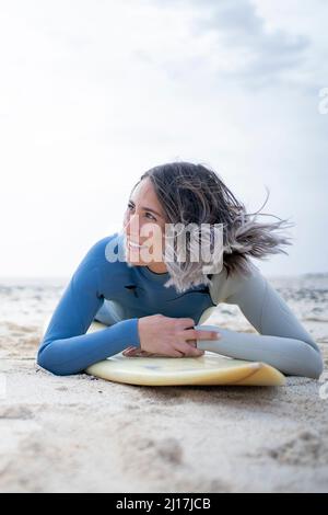 Glückliche junge Frau im Neoprenanzug, die am Strand auf dem Surfbrett liegt Stockfoto