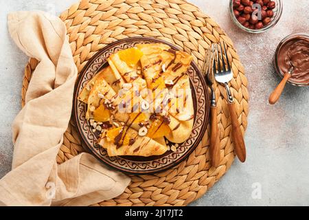 Crepes-Suzette mit Orangen und Schokolade. Dünne Crepes mit Schokoladenaufstrich, Haselnüssen und Orangenscheiben Obst auf dem Teller zum Frühstück auf altem Beton Stockfoto