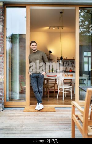Lächelnder junger Mann, der mit den Händen in den Taschen an der Tür steht Stockfoto