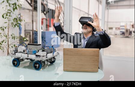 Geschäftsmann in Virtual Reality sitzend mit erhobenen Händen in der Fabrik Stockfoto