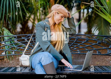 Lächelnde Frau mit langen blonden Haaren mit Laptop auf der Bank sitzend Stockfoto